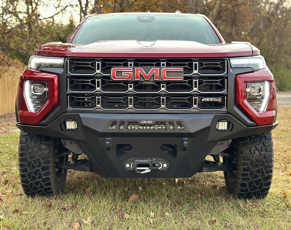 Red GMC truck with black aftermarket bumper, grille, and off-road tires, parked on grass.