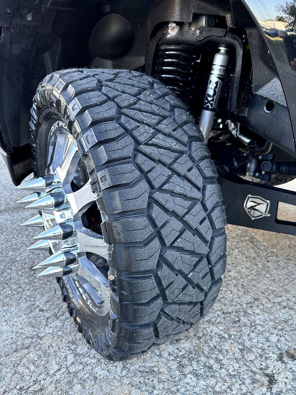 Close-up of a truck's wheel with a spiked rim and a heavily treaded tire. The truck is black.