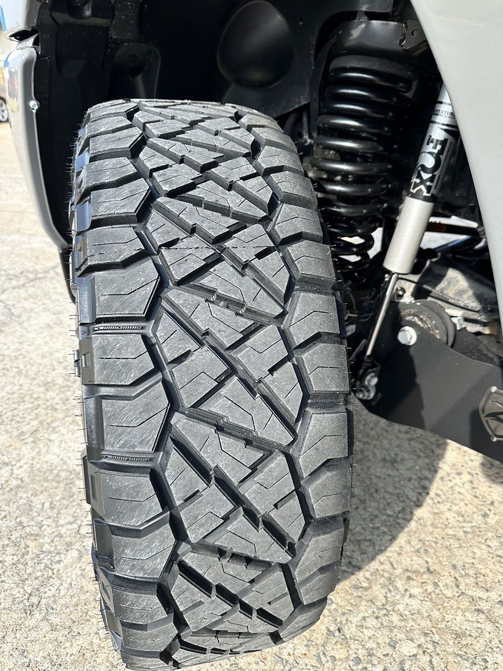 Close-up of a black off-road tire with a deep tread pattern on a vehicle with suspension components visible.
