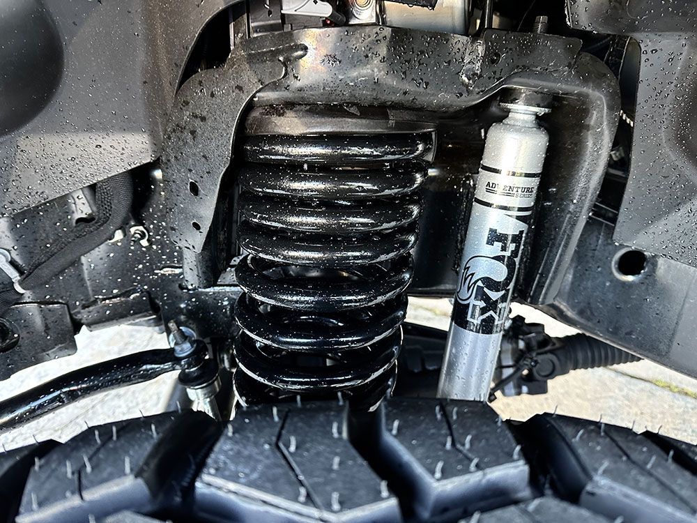 Close-up of a vehicle's suspension system. Black coil spring, silver Fox shock absorber, and a black tire are visible.