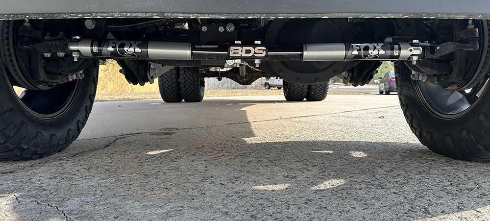 Undercarriage of a truck with a BDS steering stabilizer and dual rear wheels, parked on asphalt.