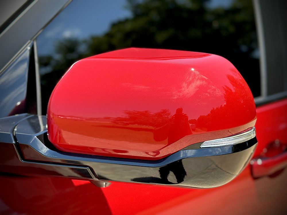 Red car side mirror, glossy finish, black trim. Reflecting sky and trees.