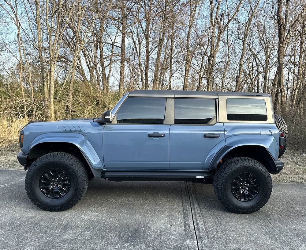 Blue Ford Bronco SUV with black wheels on a road, trees in the background.