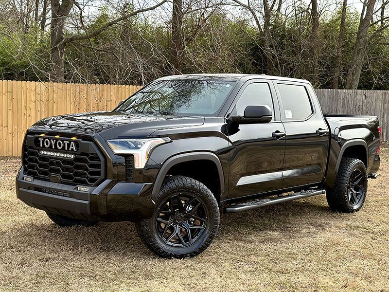 Black Toyota Tundra truck parked on grass. Black wheels, grill, and trim. Wooden fence and trees in background.