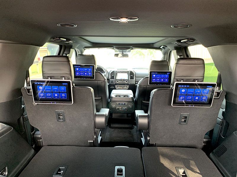 Rear view of a car interior with four headrest-mounted screens. Seats are black, and the car has a sunroof.