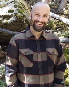 A man with a beard is wearing a plaid shirt and smiling.