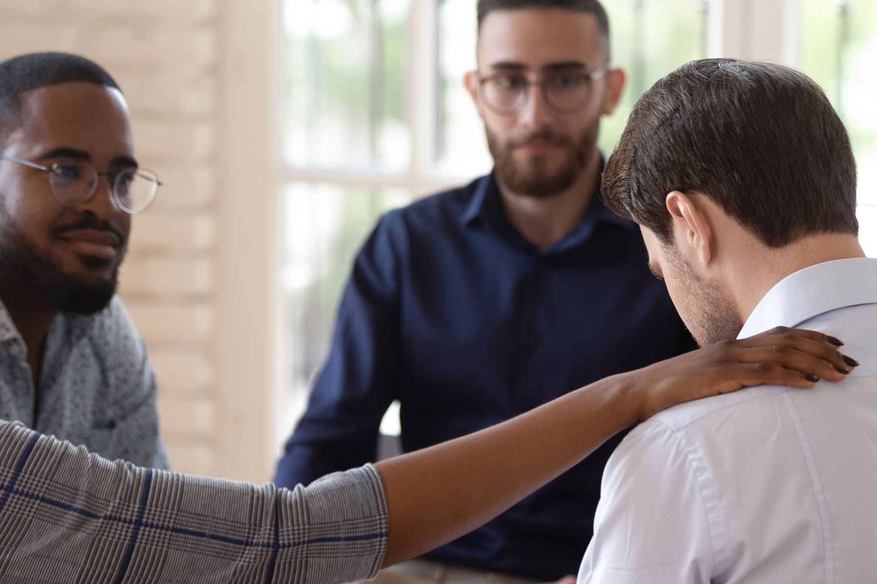 A group of men are sitting around a table and a woman is putting her hand on a man 's shoulder.