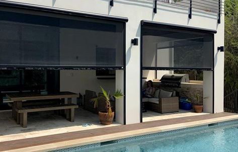 Exterior of a house with dark mesh shades lowered over an outdoor dining area and patio, near a pool.