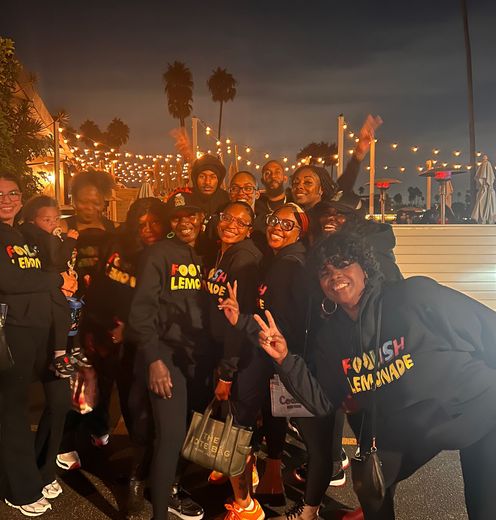 Group in black shirts posing with peace signs at a nighttime waterfront event with lights in the background