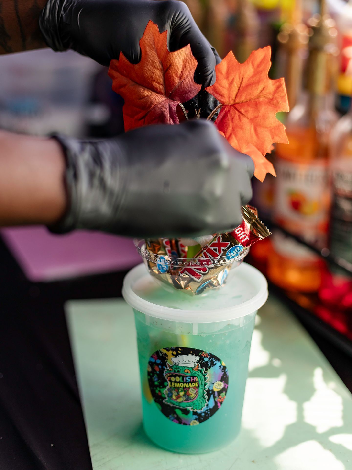Gloved hands assembling a colorful drink with orange leaves and a decorative cup lid