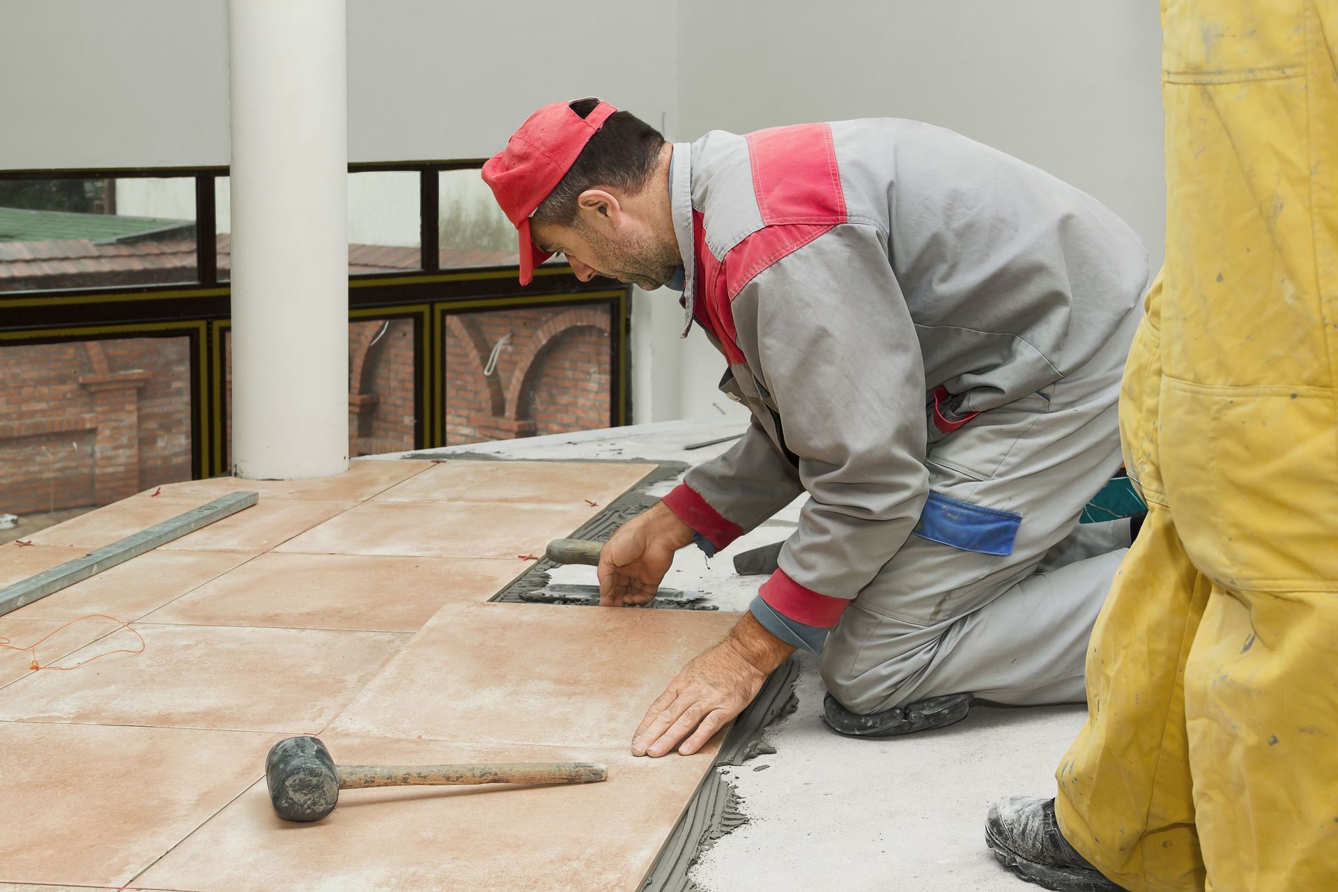 Worker kneeling on tile floor, adjusting a tile with a mallet nearby in a construction area
