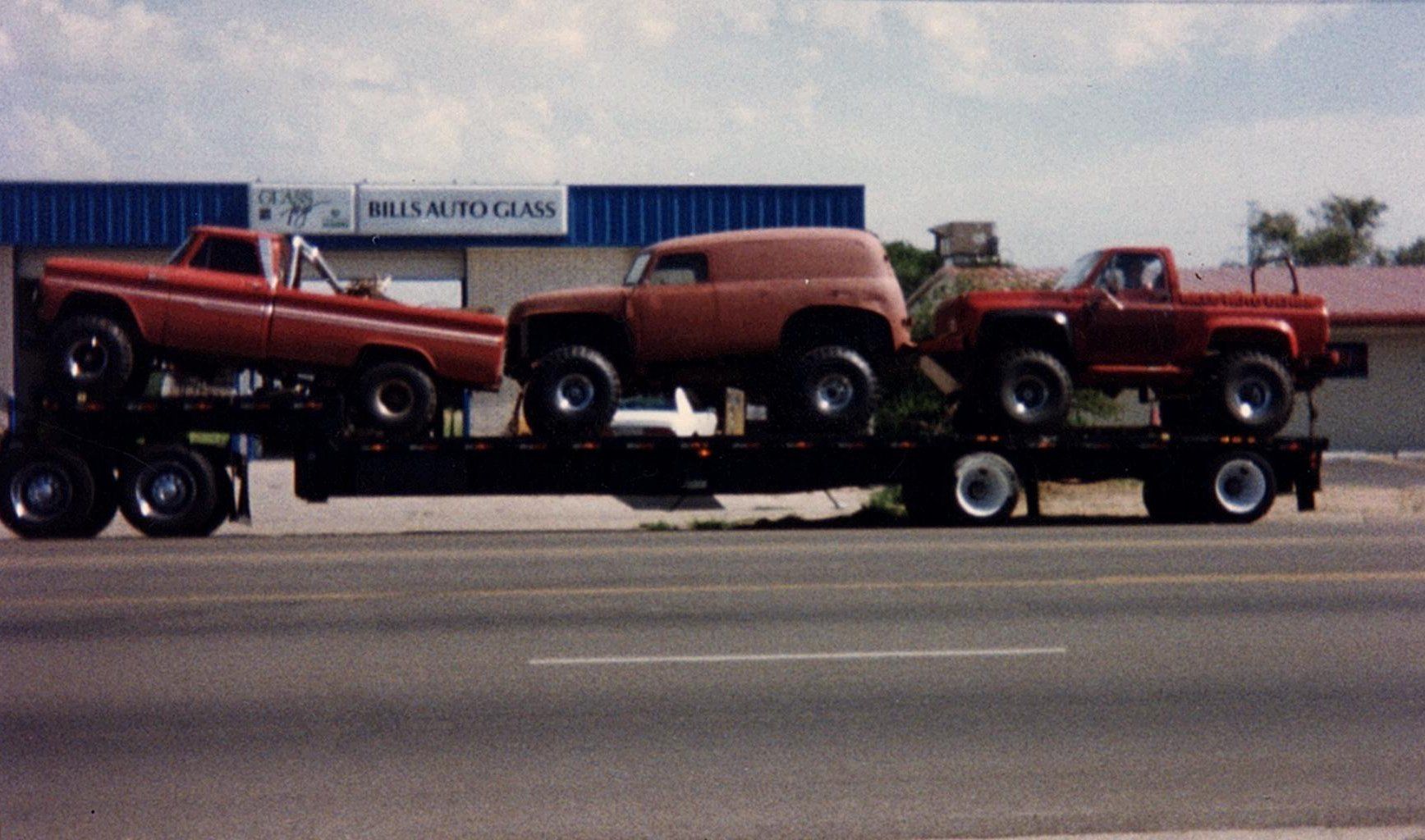 Three red trucks are on a trailer on the side of the road
