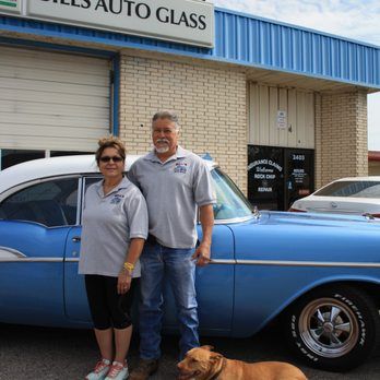 A man and woman standing next to a blue car in front of Bill's Auto Glass