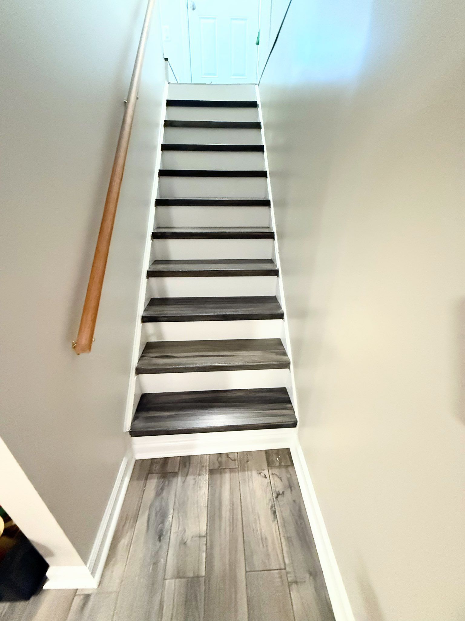 Staircase with white risers and gray and black treads. Wooden handrail on the left. The stairs lead up to a doorway.