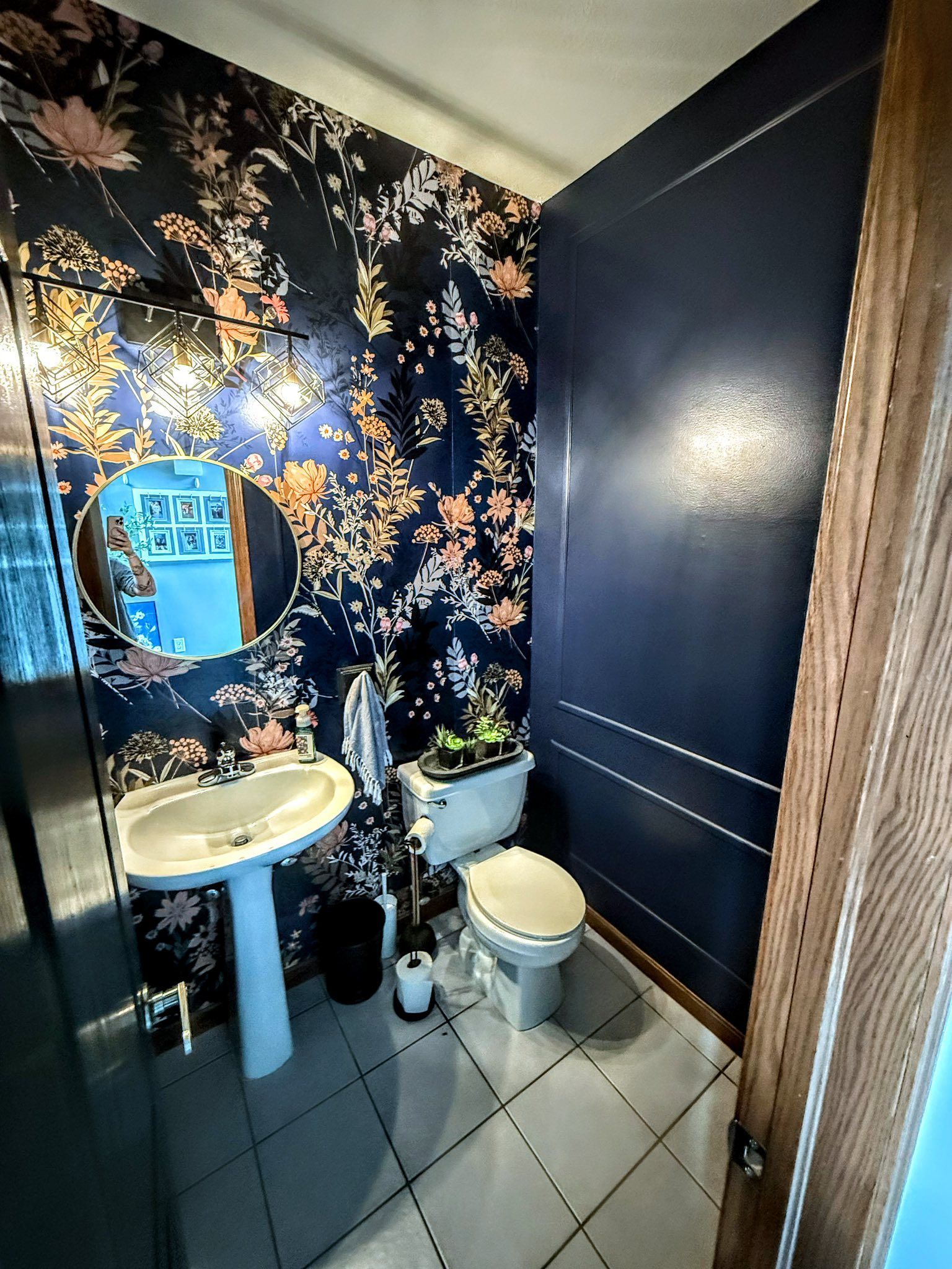 A small powder room with floral wallpaper, navy blue wainscoting, a pedestal sink, and a white toilet. A round mirror hangs above the sink.