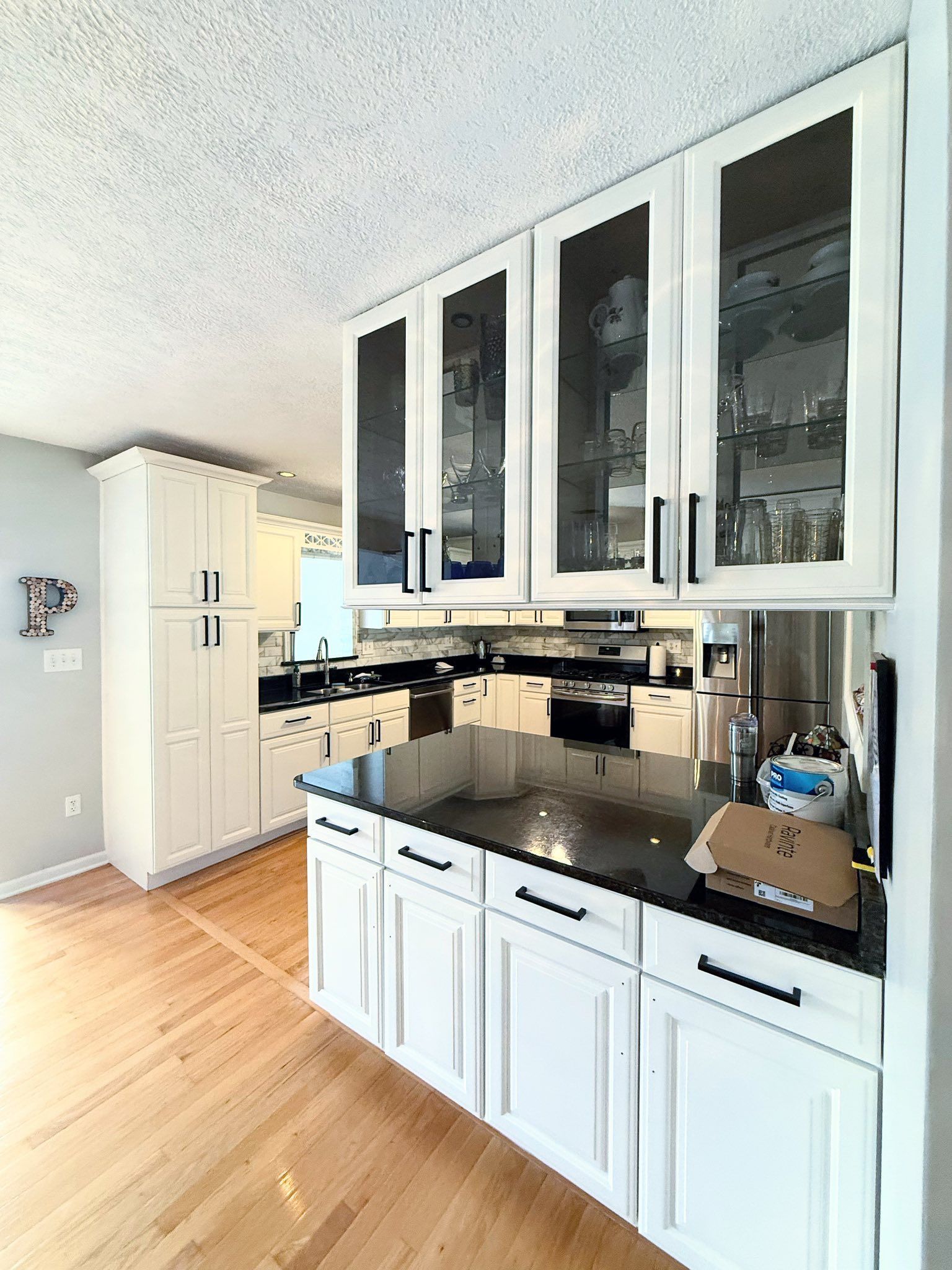 Bright white kitchen with glass-fronted cabinets, black countertops, and hardwood floors. There is a large island and matching cabinets.