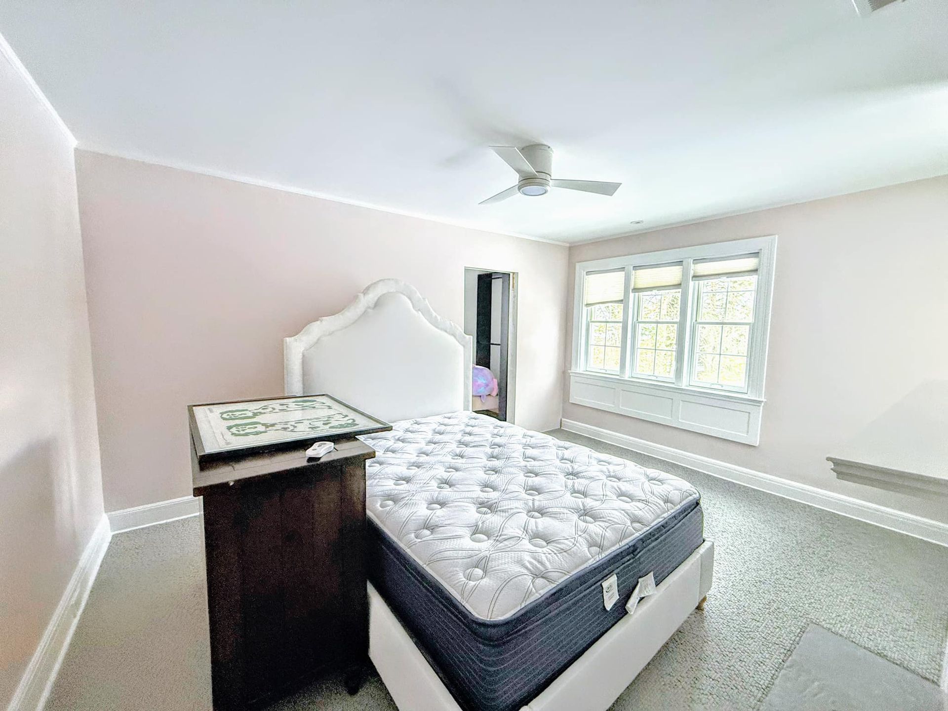 A bedroom with a white bed, a dark nightstand with a framed picture, and a window. The walls are painted light pink.