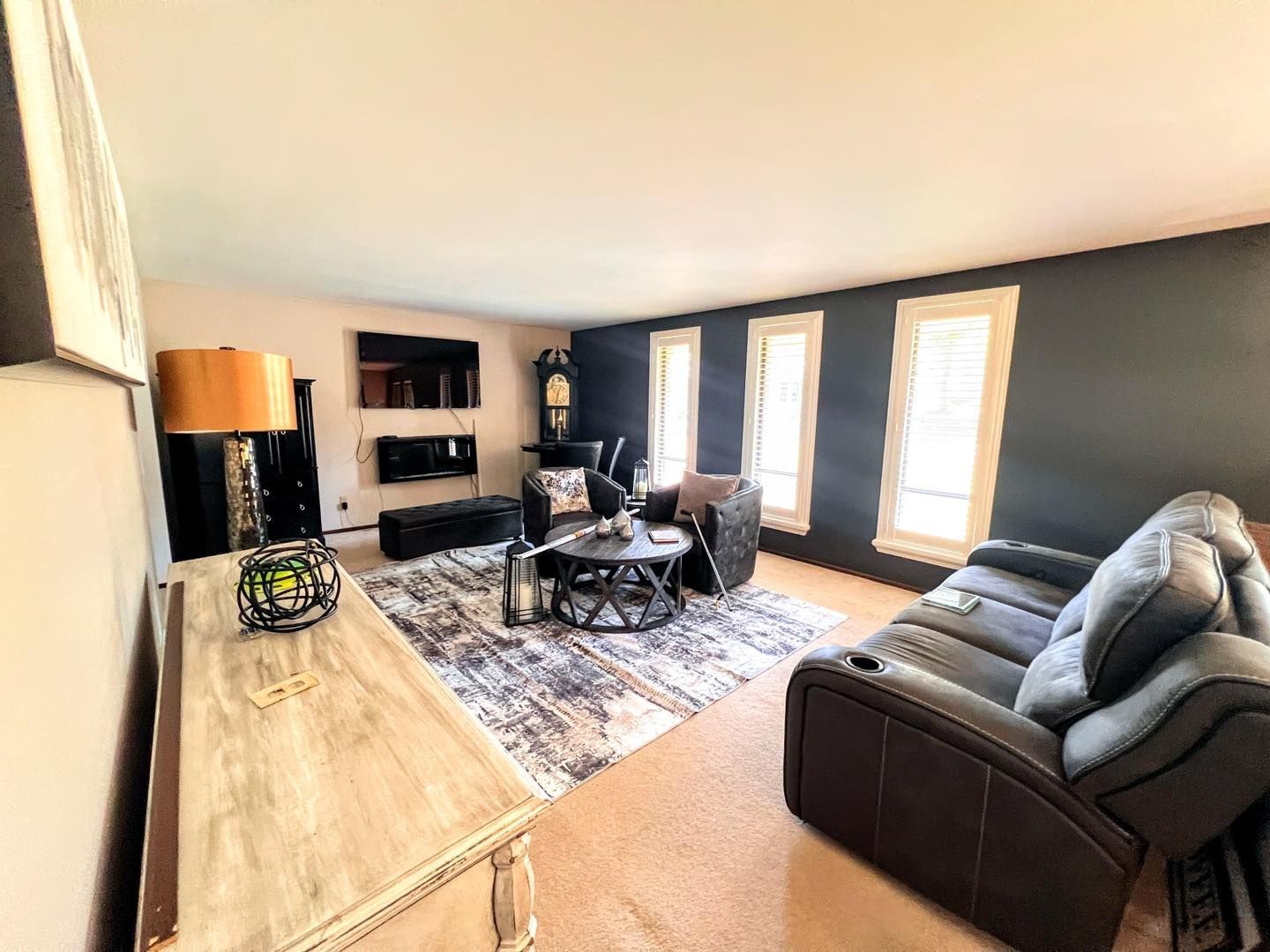Living room with dark accent wall, fireplace, and brown reclining sofa. Beige rug, light wood console, and large windows.