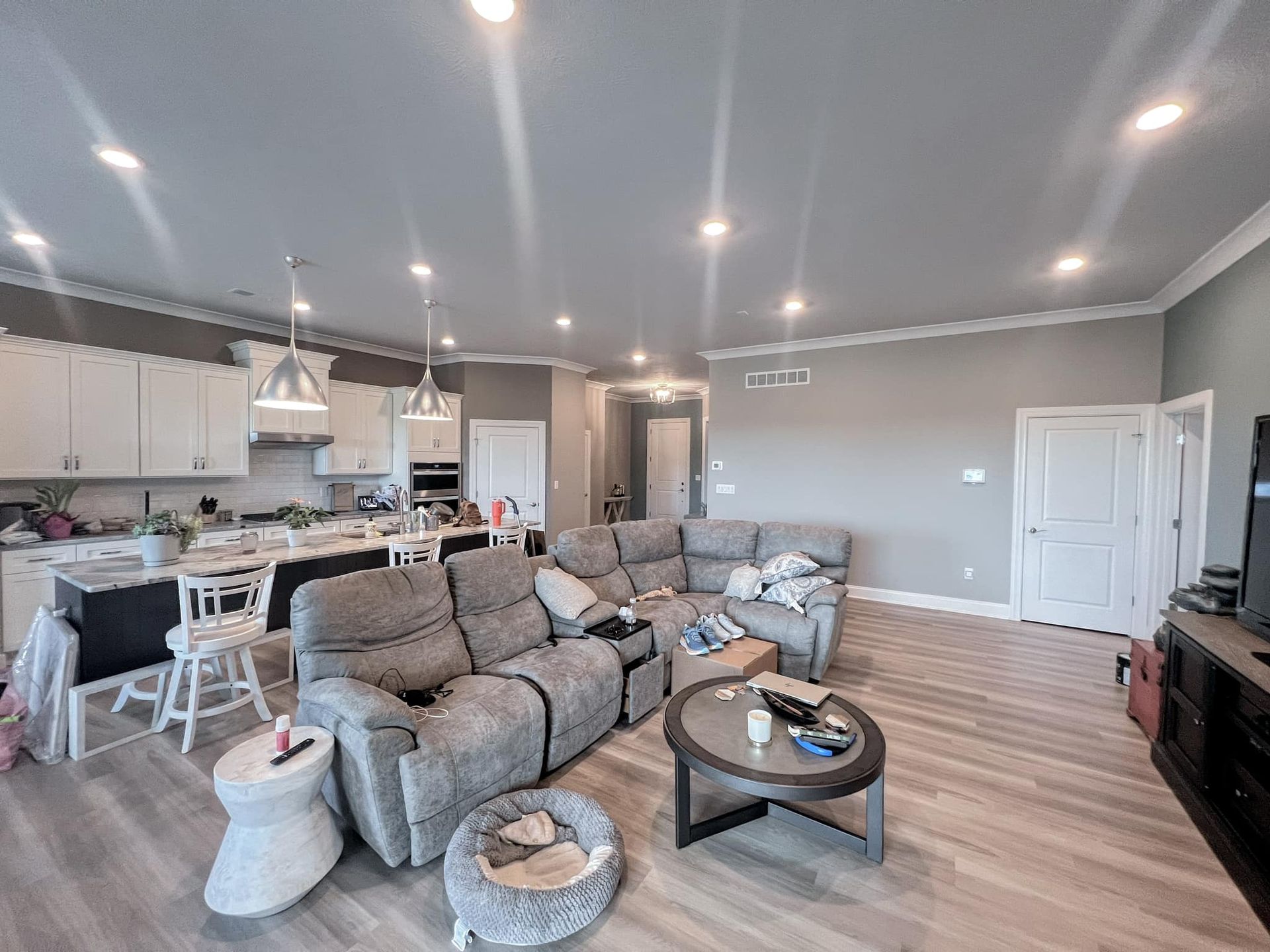 Spacious living room with an open kitchen; features a gray sectional sofa, round coffee table, and a light-colored hardwood floor.