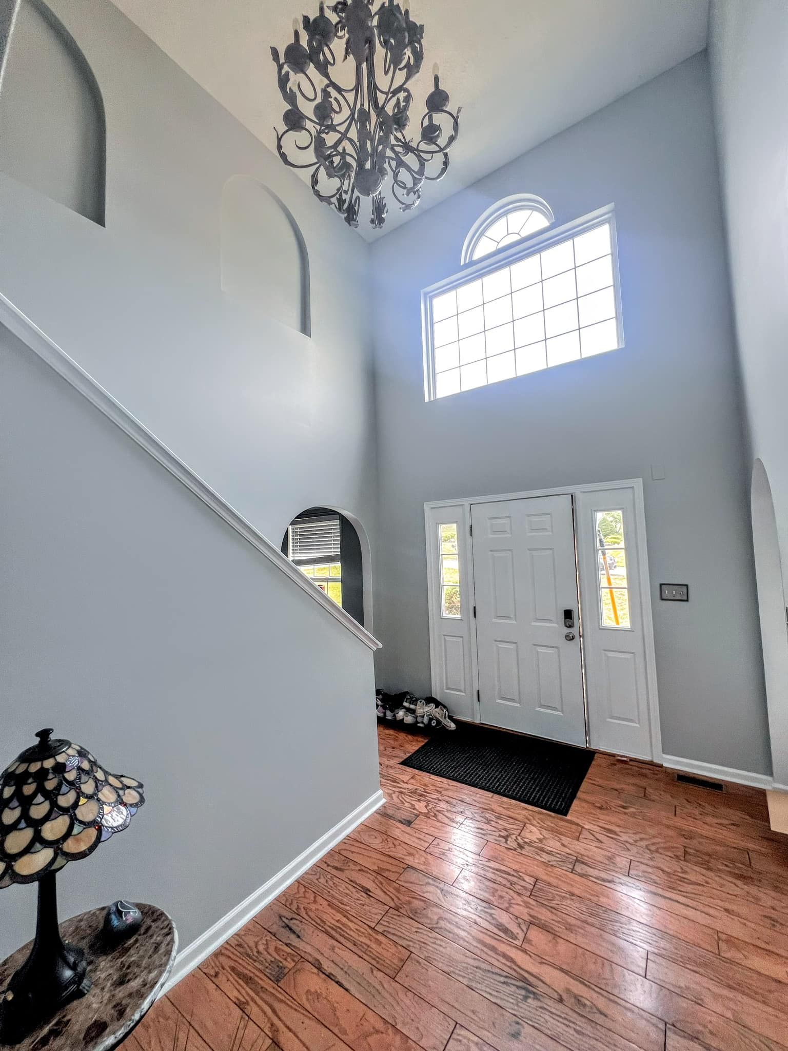 A bright entryway with light blue walls and a dark wood floor. A chandelier hangs above the white double doors and a staircase.