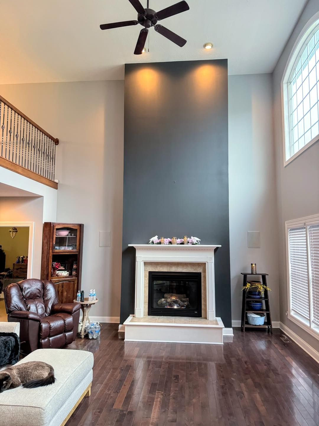 A spacious living room with a fireplace centered on a tall gray wall. Dark hardwood floors, a cat on the couch, and a balcony are visible.