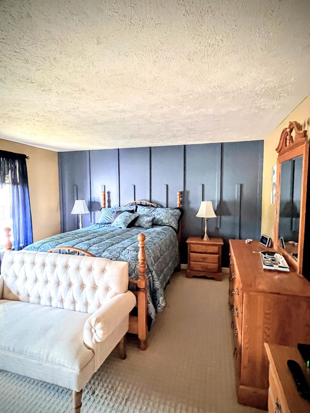 Bedroom with a blue accent wall behind a wooden bed, a white loveseat, and wooden dresser and nightstand.