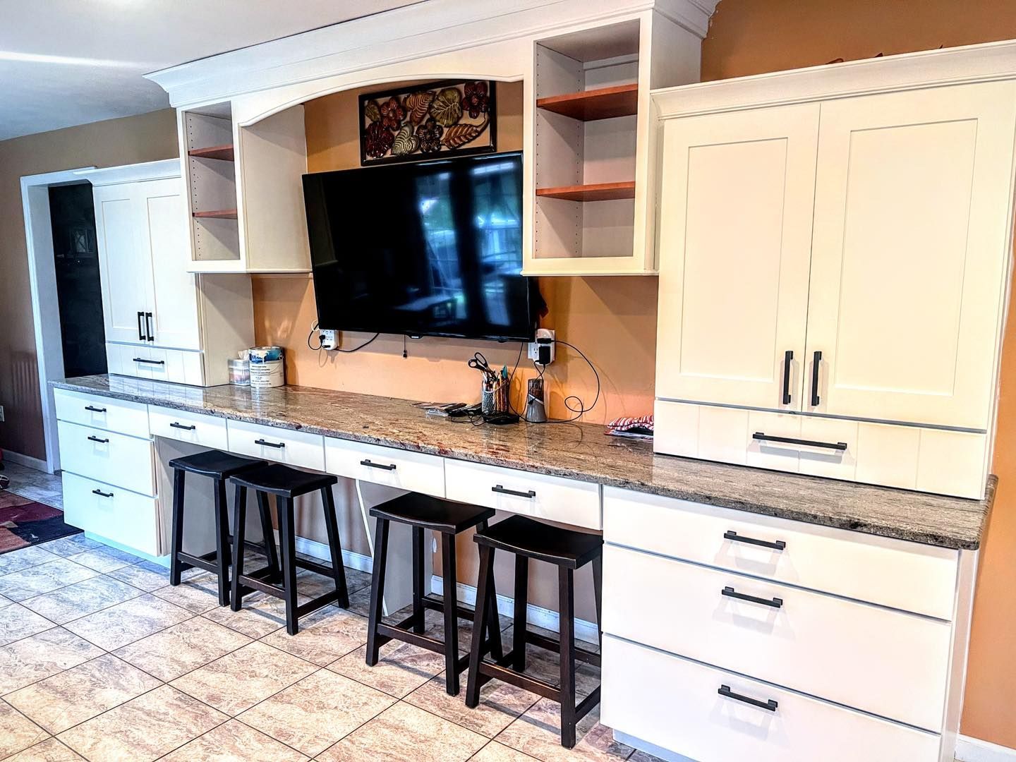 Built-in kitchen desk with white cabinets, a granite countertop, and black stools. A TV is mounted above the desk.