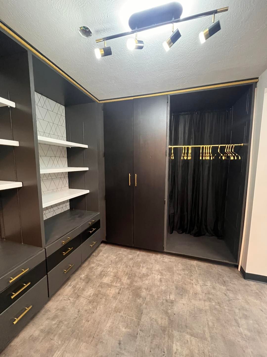 Walk-in closet with dark gray cabinetry and gold accents. Features shelves, drawers, a wardrobe, and a curtained hanging space.