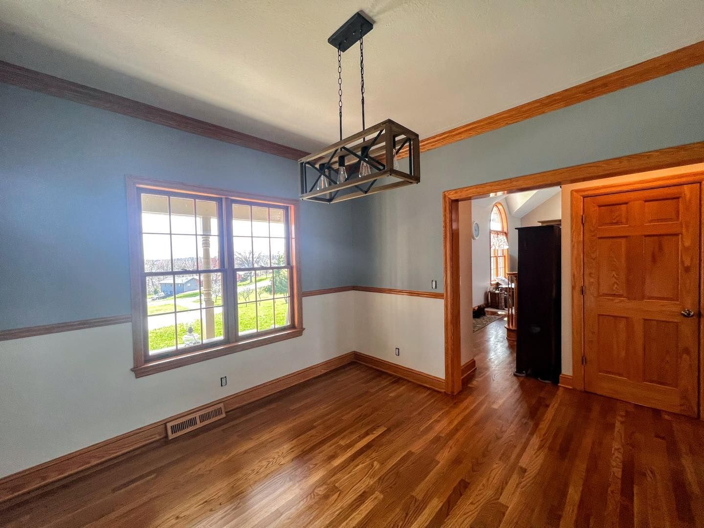 Dining room with wood floors, trim, and a blue-and-white wall. A rectangular chandelier hangs above the space, and a view from the window is seen.