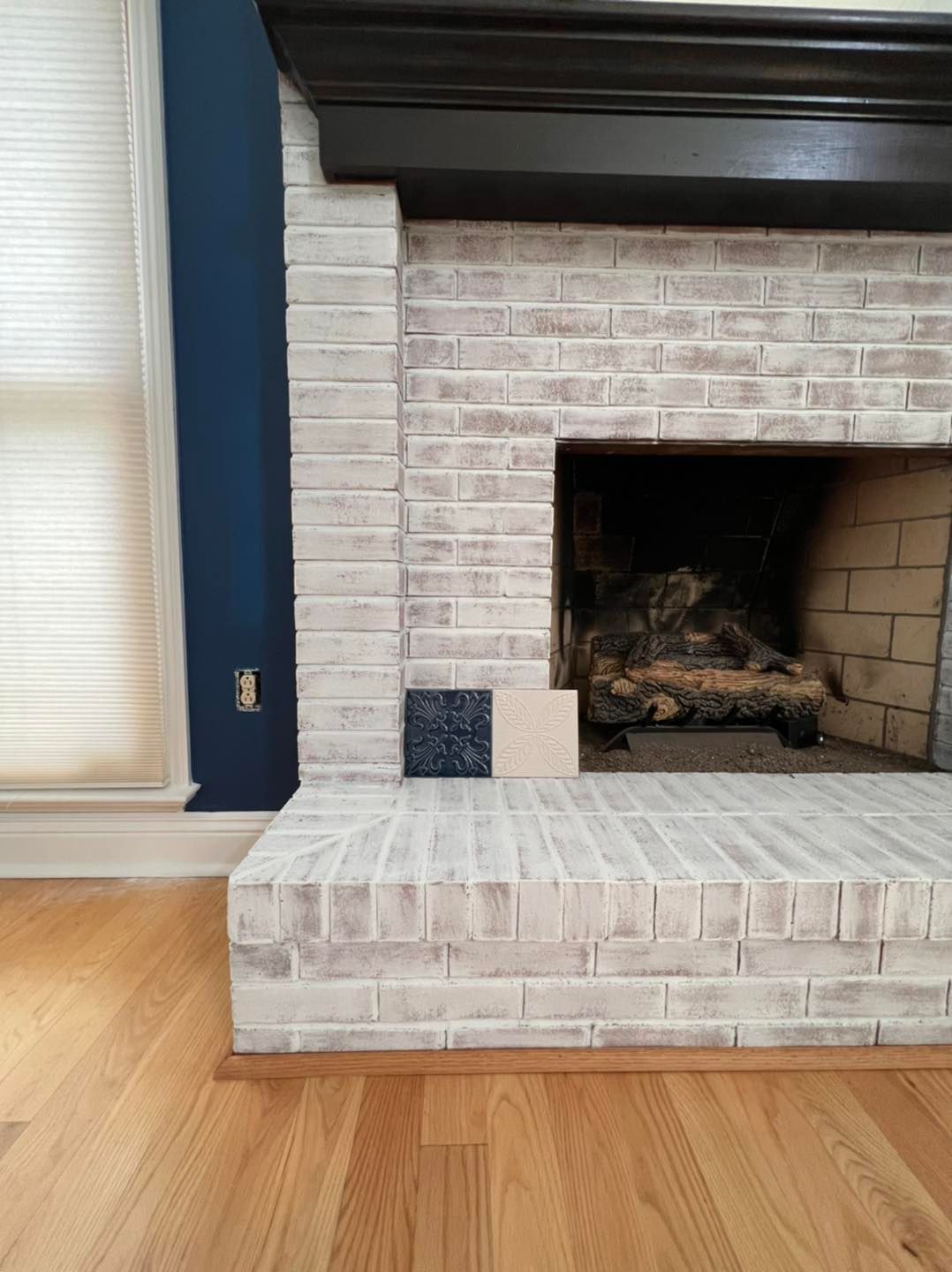 A whitewashed brick fireplace with a black mantel, blue wall, and wood flooring. Samples of dark and light tile are placed on the hearth.