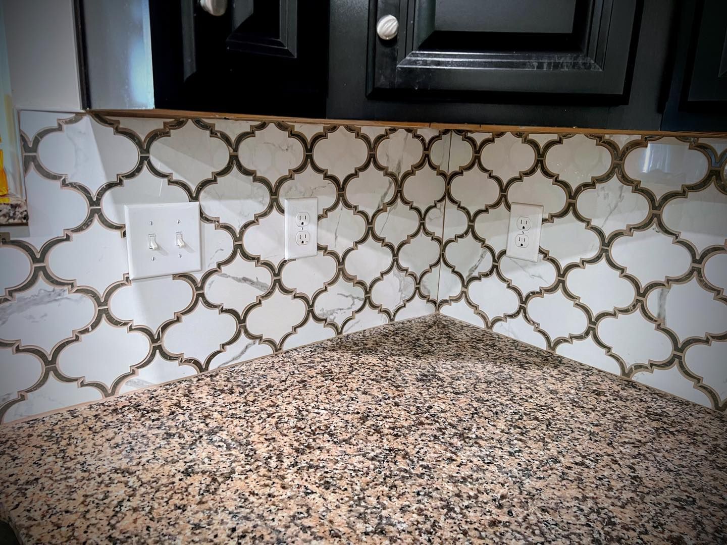 Kitchen backsplash with white and gold patterned tiles installed above a granite countertop with two electrical outlets.