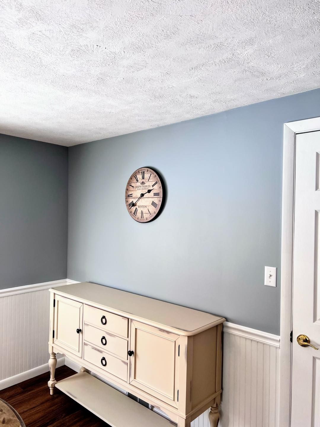 A blue-painted room with a cream-colored buffet, a round clock, and a white door. The lower walls have white beadboard.