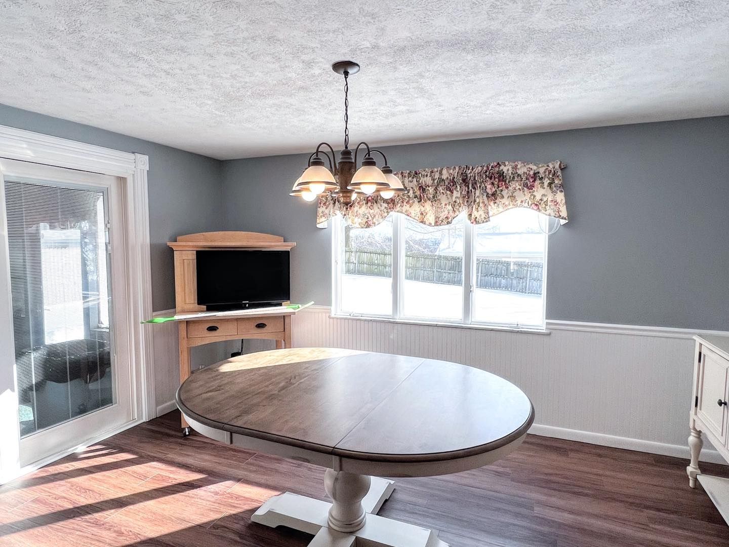 A dining room with a round table, TV console, and a window with a floral valance; sunlight streams in.