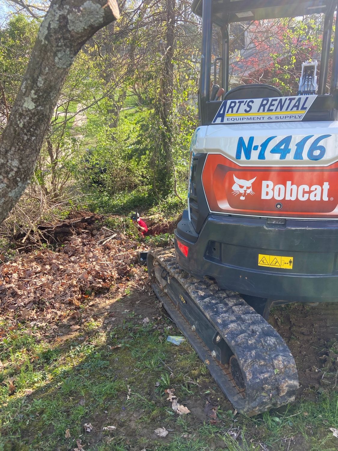 A bobcat excavator is parked in the grass next to a tree