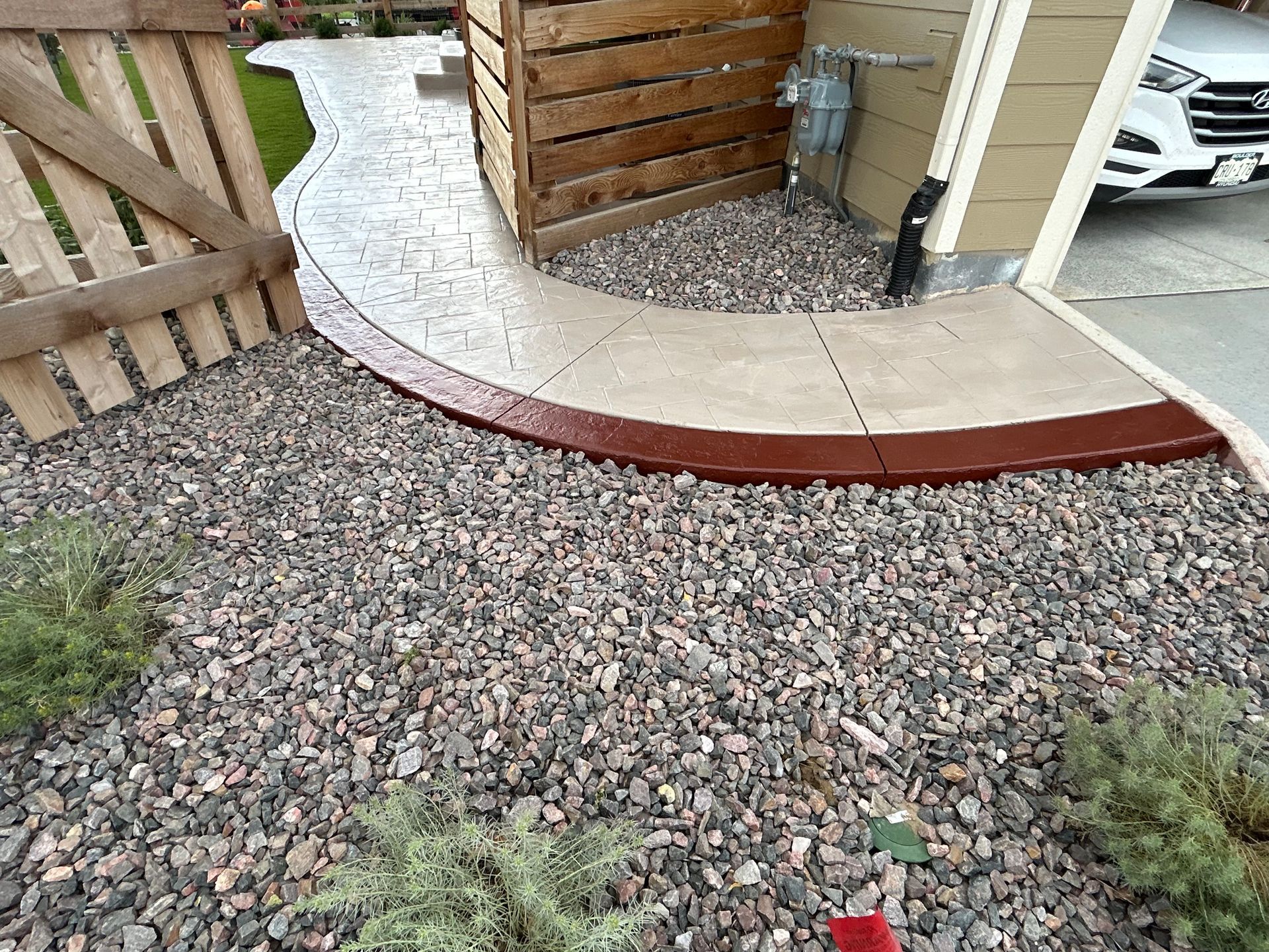 A concrete walkway with a red border is surrounded by gravel and a wooden fence.