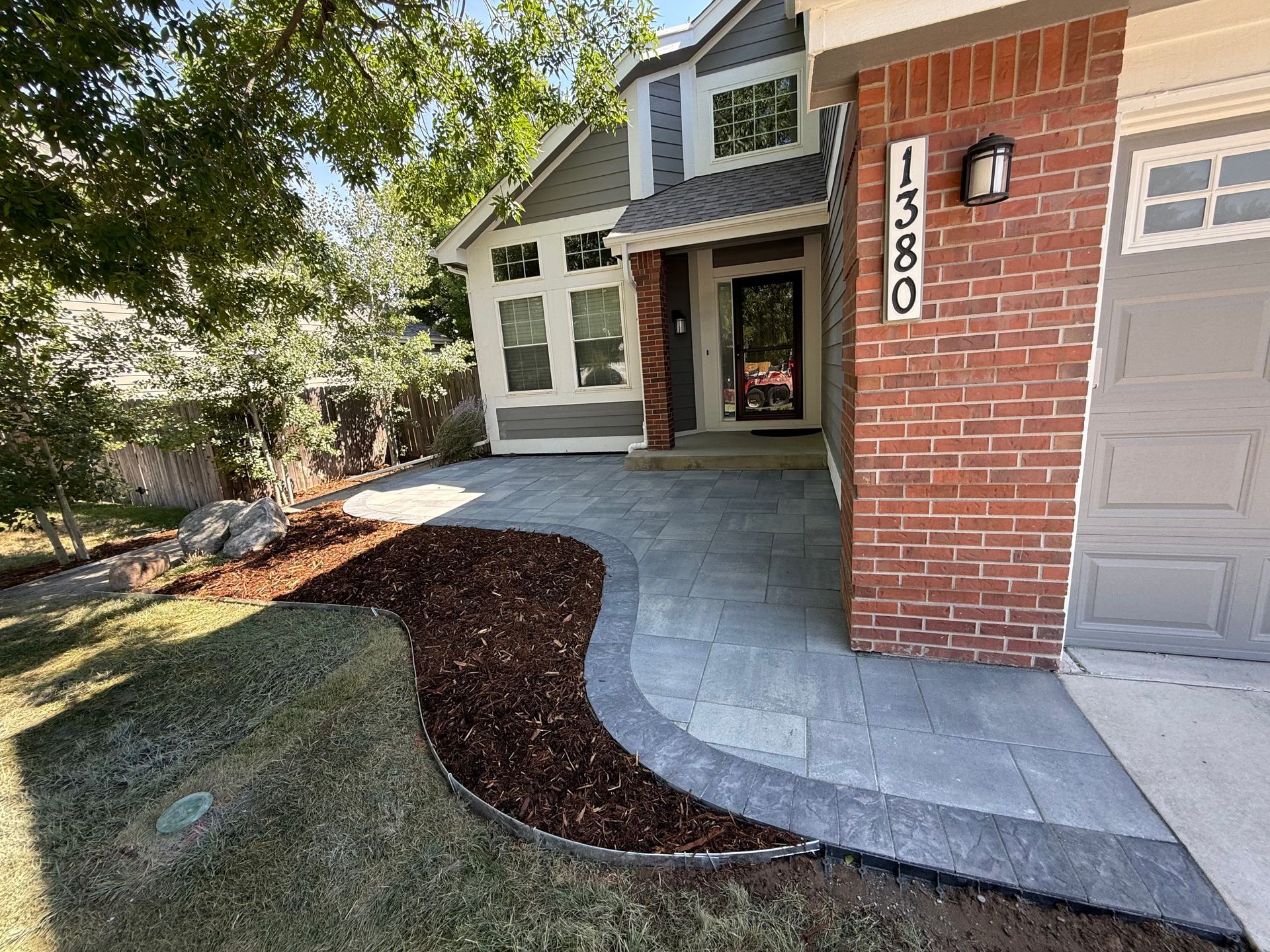 A house exterior with a new paver walkway, dark mulch flower bed, brick facade, and a grey garage door.