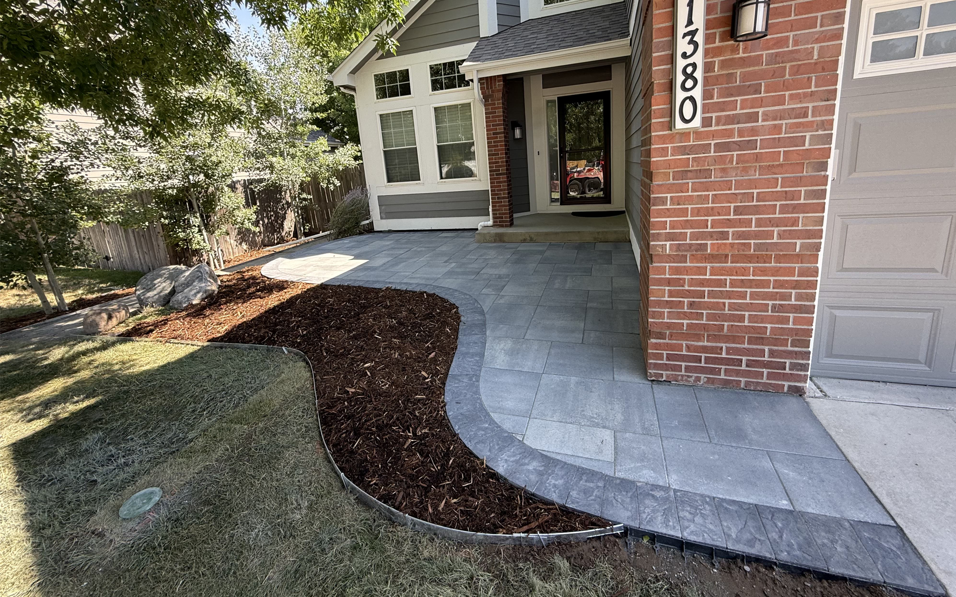 A new stone paver walkway leads to the front door of a brick house, bordered by a fresh mulch garden bed.