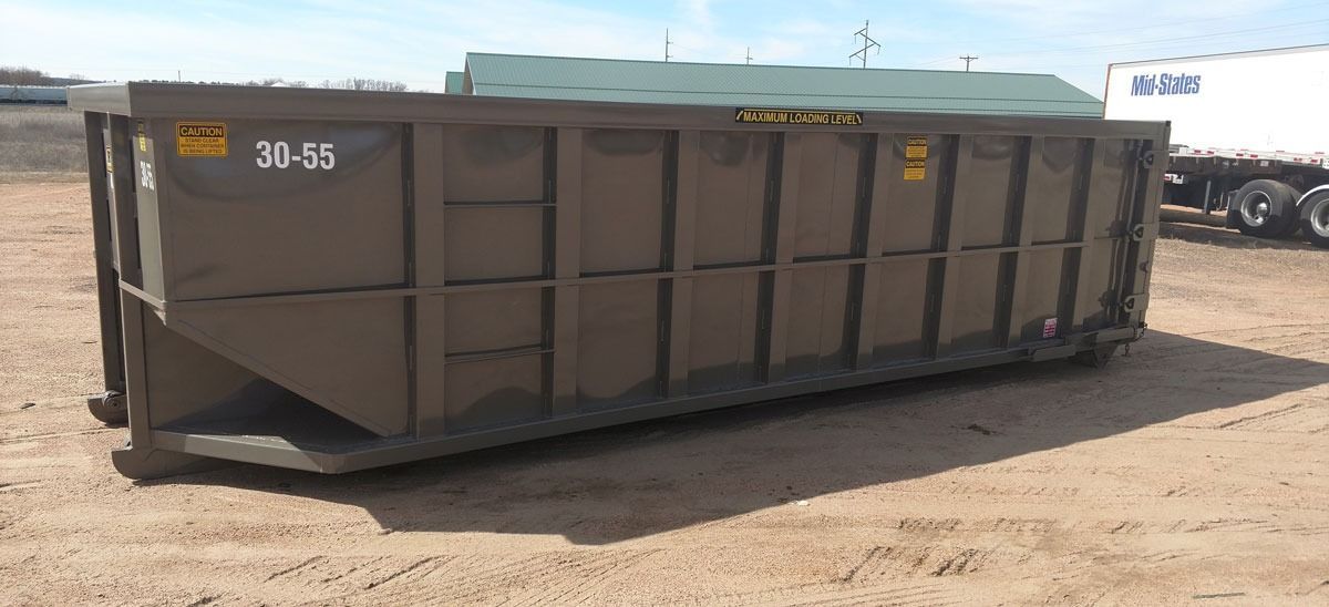 A large, brown dumpster sits on a sandy, outdoor area. A trailer truck is visible in the background.