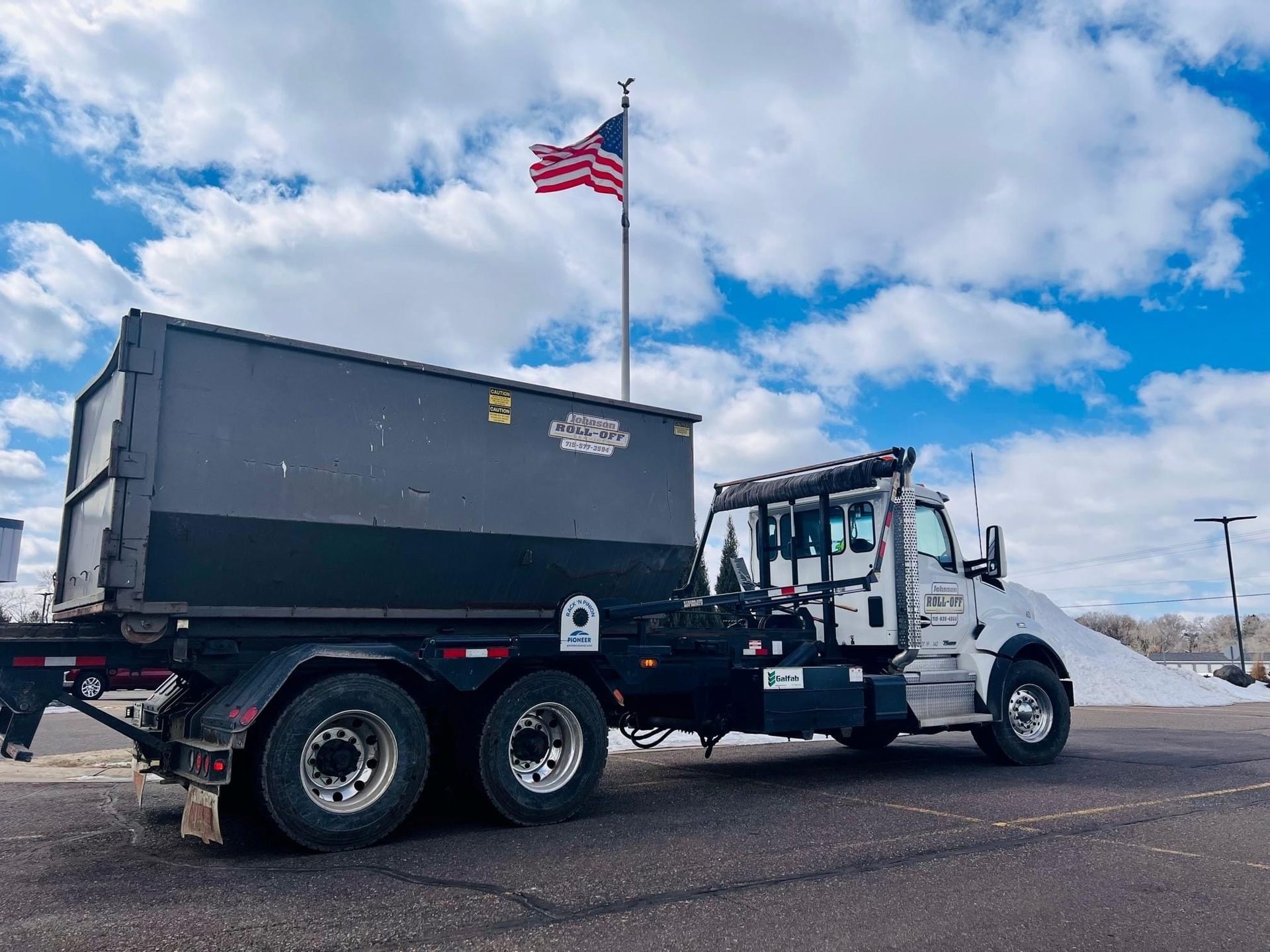 Dump truck with large container, parked outside under a cloudy sky with American flag.