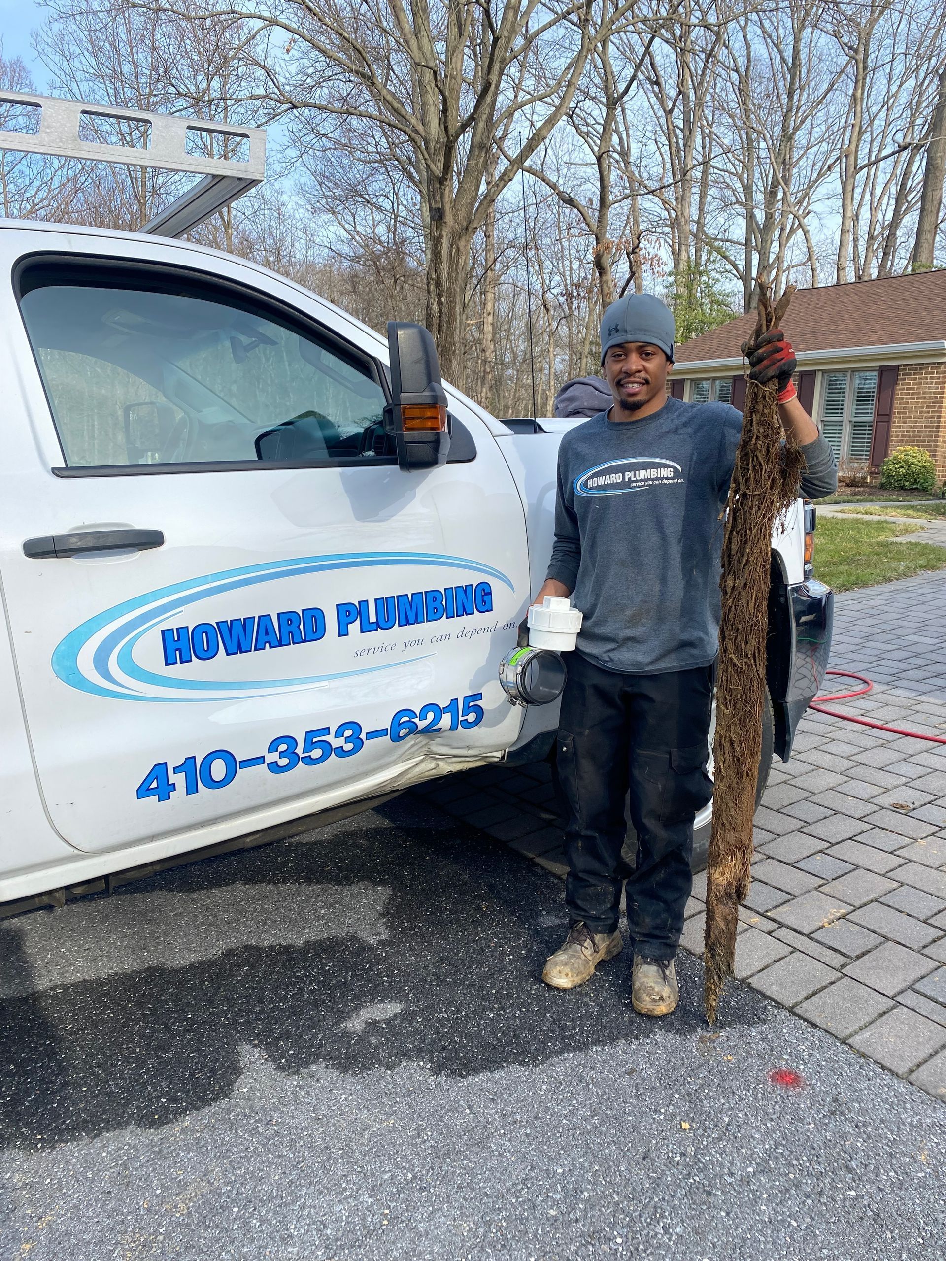 A smiling plumber stands next to their van holding a long mass of tree roots removed from a sewer line.