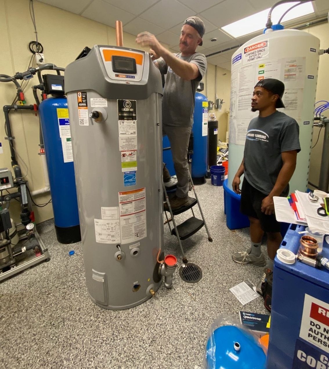 Two technicians work on a large water heater in a utility room with tanks and tools.