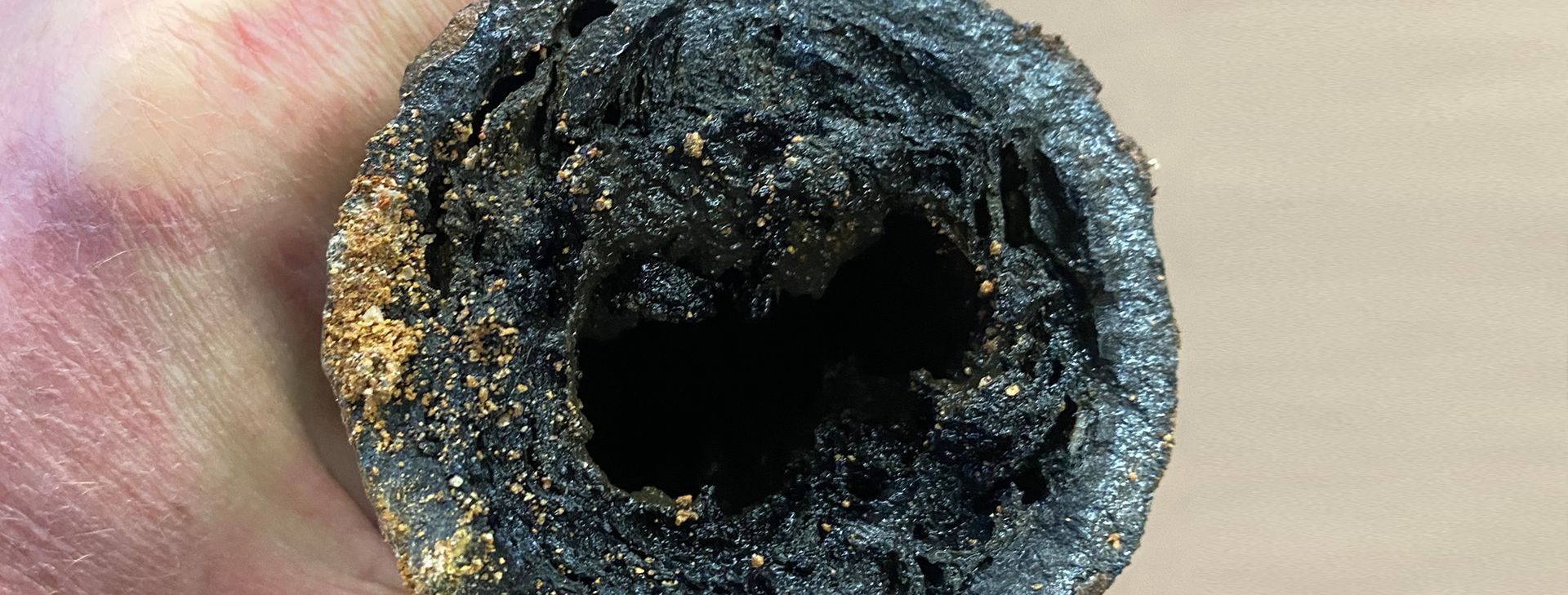 A hand holds a pipe cross-section heavily clogged with thick, dark, textured sludge, leaving only a small central opening.