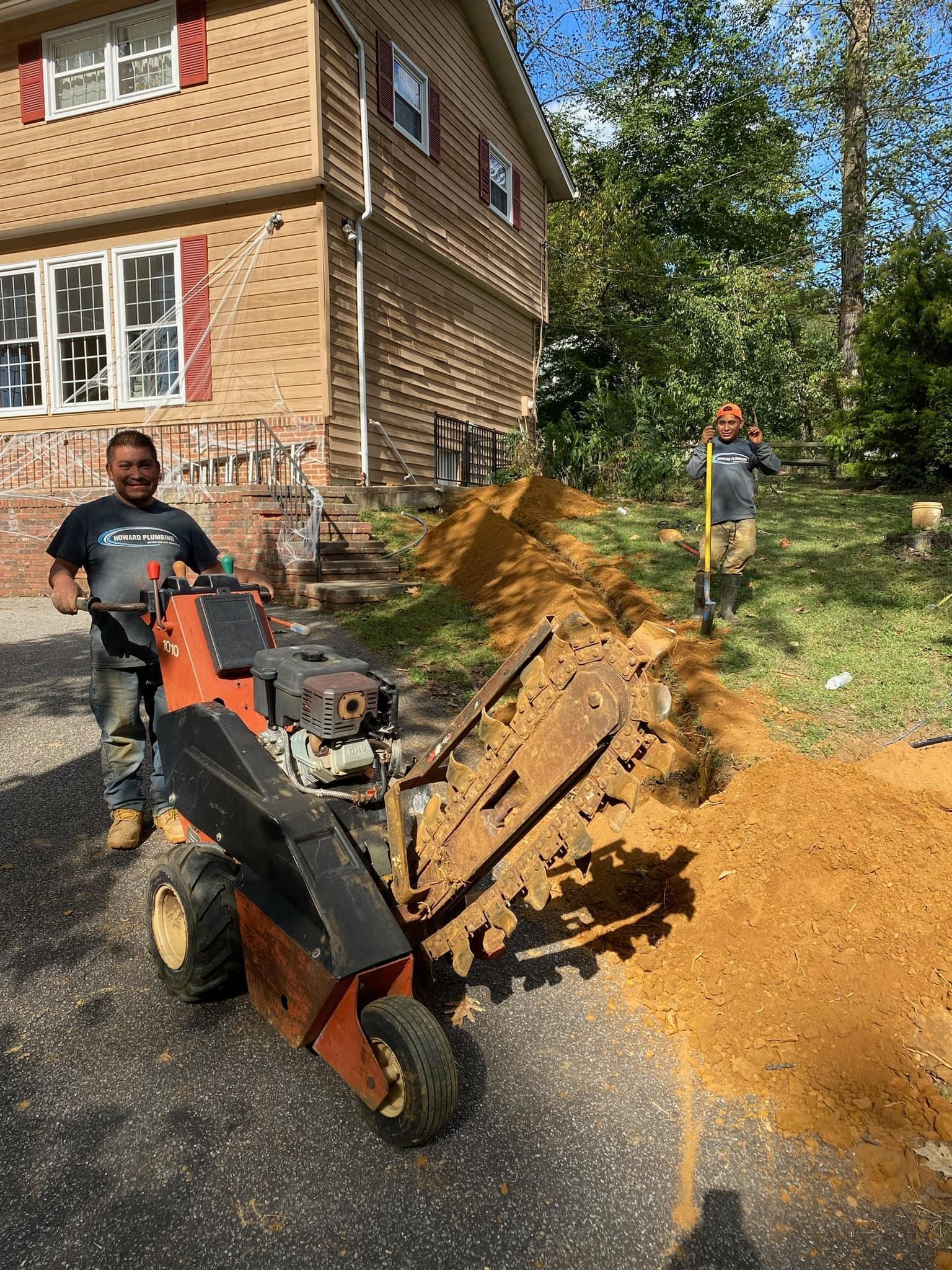 Two workers operate a trencher machine next to a house with an excavated dirt trench in the yard.