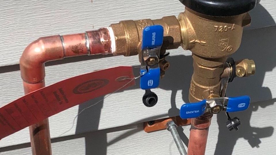 A brass backflow prevention assembly with blue-handled ball valves connected to copper piping on a wall.