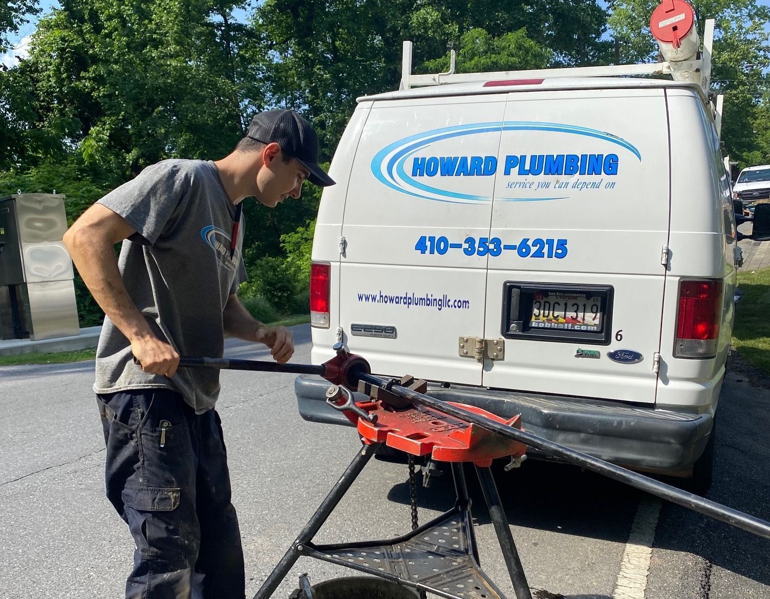 A worker uses a tripod-mounted pipe cutter to work on a long piece of pipe outside a white Howard Plumbing service van.