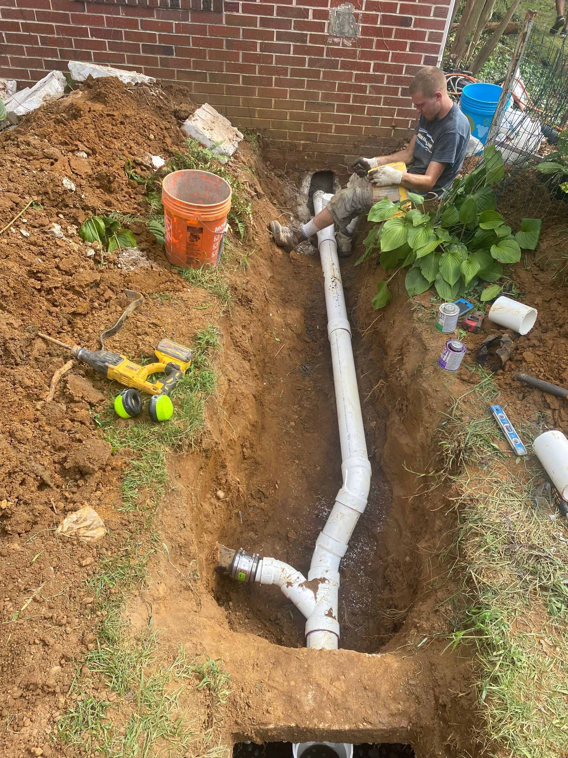 A person in a trench by a brick wall installs white PVC sewer piping connected to an existing pipe.