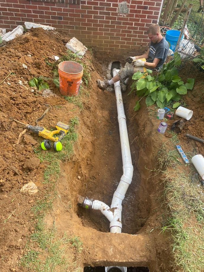 A person in a trench by a brick wall installs white PVC sewer piping connected to an existing pipe.
