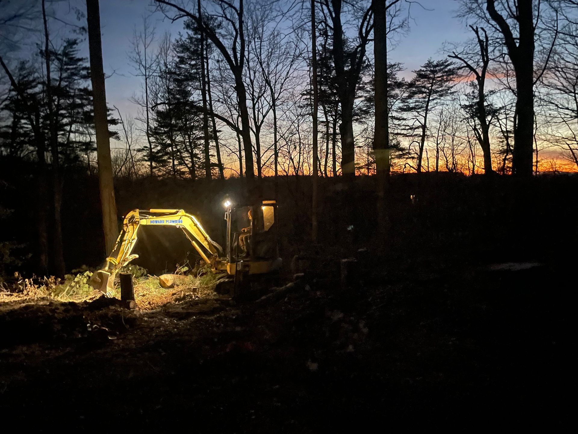 A yellow excavator working in a wooded area during a colorful sunset, with its work lights on.