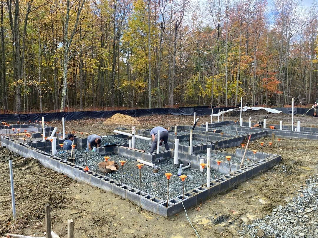 Construction workers install plumbing pipes and concrete blocks for a residential building foundation in a wooded area.