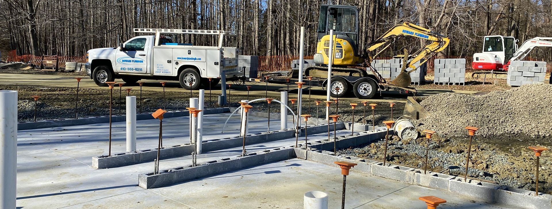 Construction site with a white utility truck, yellow excavator, and concrete foundation forms on a gravel lot.